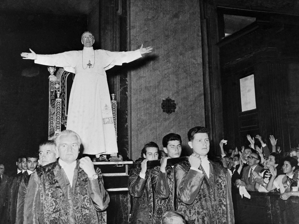This unusual photo shows Pope Pius XII standing before his gestatorial chair as he is carried into one of the Vatican's apostolic halls to bless crowds gathered there. Pilgrims in great numbers are presently crowding the Vatican to be blessed before the pontiff retires to his summer palace shortly.