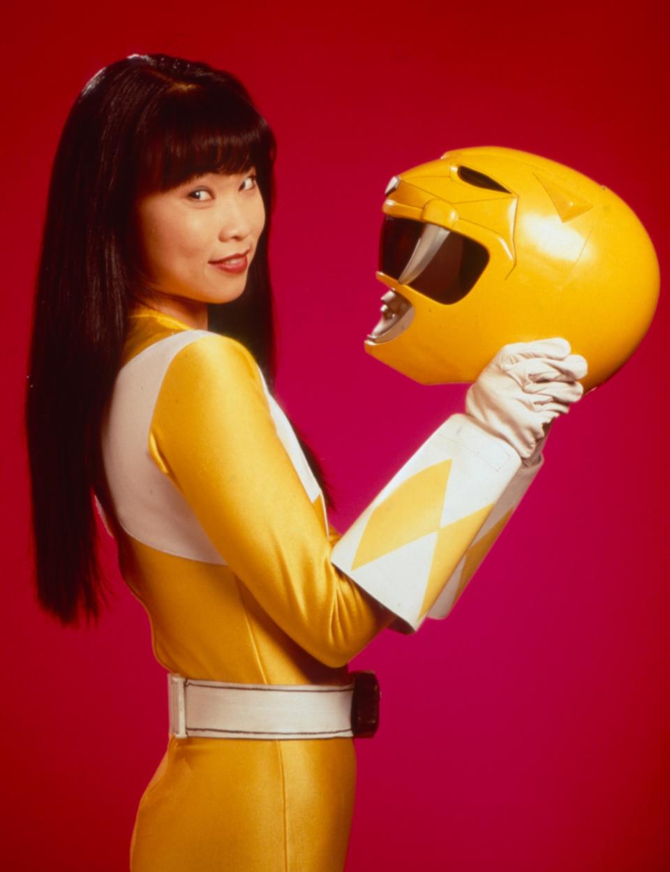 Thuy Trang in a yellow and white Power Rangers suit, holding a yellow helmet