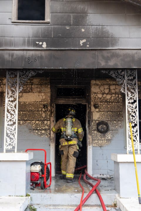 Firefighter entering a white wood building. There are visible burn marks on the side of the house.