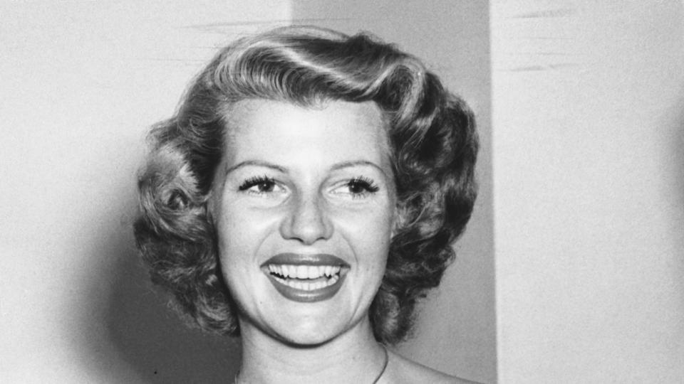 rita hayworth smiles and looks right of the camera, she clasps her hands in front of her chest and wears a black dress with a bow in the front and a medallion necklace
