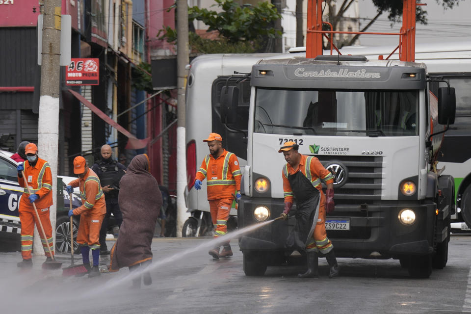 A homeless drug user walks past city workers cleaning the streets in the Santa Efigenia shopping district, a major commercial hub for electronics, technology products, and accessories in downtown Sao Paulo, Brazil, Thursday, May 18, 2023. The decline of Sao Paulo's downtown area has accelerated over the last year, where crack users seem to be everywhere, roaming the central streets of South America's biggest city. (AP Photo/Andre Penner)