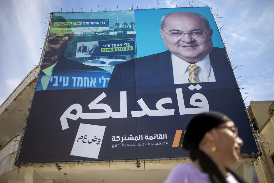 In this Thursday, March 5, 2020 photo, Israeli Arab Fadila Maha walks past an election campaign poster showing Israeli Politician Ahmad Tibi of the Joint List in Tira, Israel. A surge in Arab voter turnout was key to depriving Prime Minister Benjamin Netanyahu and his nationalist allies of a parliamentary majority in this week’s Israeli election. Undercutting Netanyahu’s ambitions was celebrated as sweet payback in the nearly 2 million-strong minority that the hard-line leader had relentlessly tried to tarnish as disloyal to the state. The Arabic reads "I sit with you." (AP Photo/Ariel Schalit)
