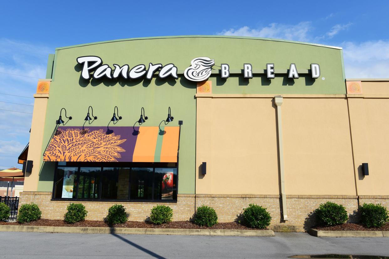 Exterior of Panera Bread bakery in Johnson City, Tennessee surrounded by shrubbery and parking spots with outdoor seating area on the left with a blue sky in the background