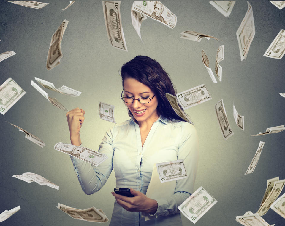 A woman checking her smartphone and pumping her fist in excitement as cash money falls around her.