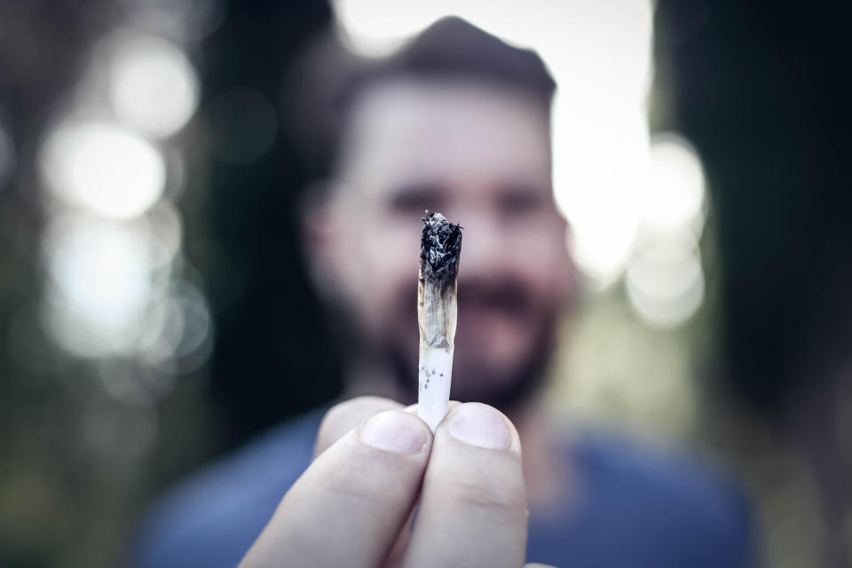 A bearded man holding a lit cannabis joint in his outstretched fingertips.