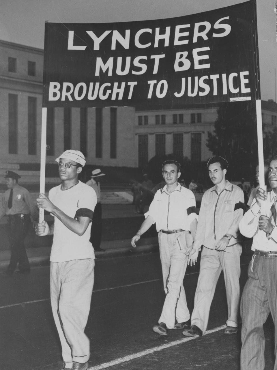 White and African American citizens march wearing black armbands and holding a sign readings 'Lynchers Must Be Brought to Justice' while police look on, Washington DC, 1946.