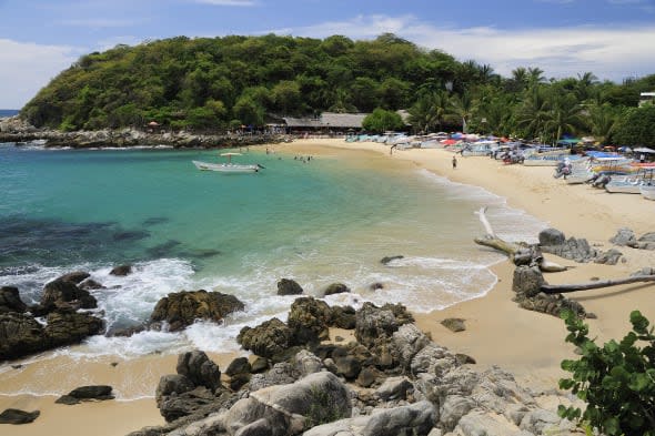 Mexico, Oaxaca, Puerto Escondido, View onto Playa Manzanillo beach with rocks and driftwood in foreground, tourist boats, people