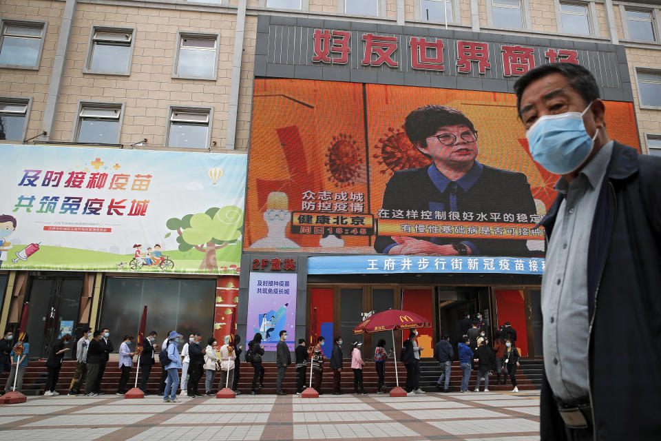 A man wearing a face mask to help curb the spread of the coronavirus walks by masked residents lining up for COVID-19 vaccine at a coronavirus vaccination site with a board displaying the slogan, "Timely vaccination to build the Great Wall of Immunity together" in Beijing, Wednesday, April 21, 2021. (AP Photo/Andy Wong)