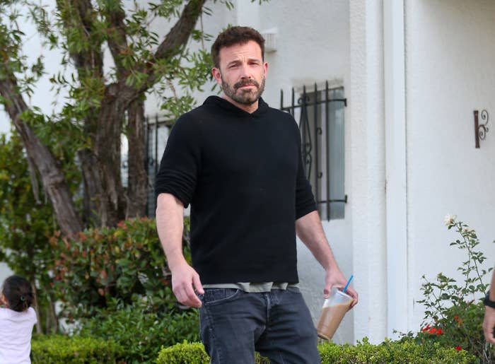 Ben Affleck is seen casually dressed in a black hoodie and jeans, holding a drink, walking outside near a white building and greenery