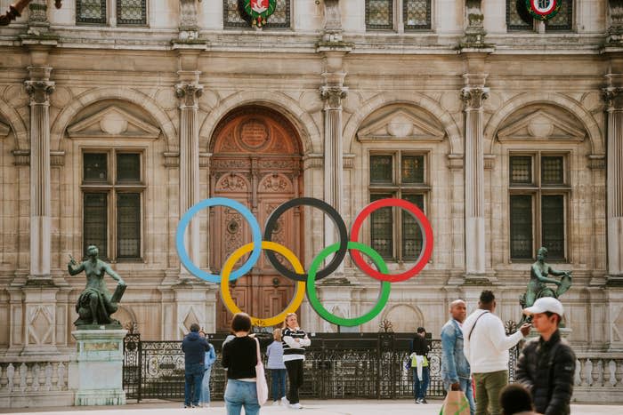 People are seen in front of a building with large Olympic rings displayed on it. The image depicts a sports-related scene. Names of specific individuals are not provided