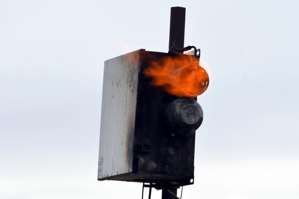 charred metal box with flames coming out of a tube protruding from the side against a white cloudy sky