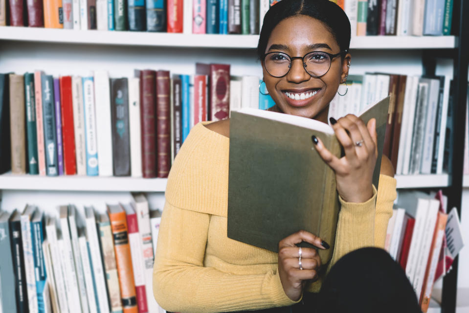 Positive african american young woman in eyeglasses for vision correction laughing while looking away and holding book in hands.Cheerful dark skinned student enjoying literature plot from bestseller