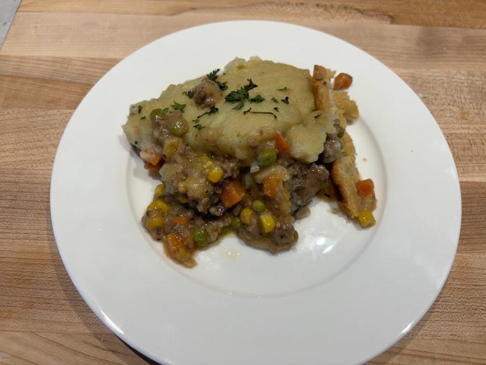 A slice of shepherd's pie with pieces of beef, carrots, corn, peas, and mashed potatoes on a white plate on a cutting board