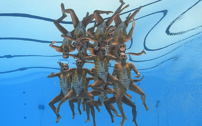 The Mexico swim team competes in artistic swimming. The camera has captured the group of swimmers completely submerged, and the surface of the water reflects the group of swimmers. Their arms and legs are bent as they stay suspended below the surface
