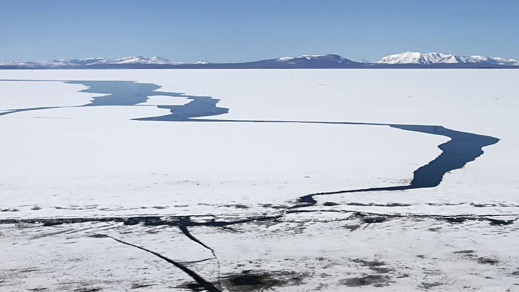  A frozen lake landscape with large cracks in the ice showing the deep blue water beneath. There are snow-capped mountains in the distance. 