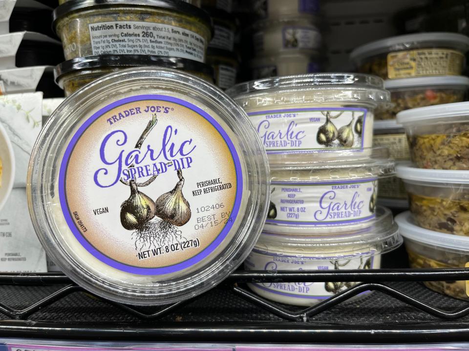 A black shelf at Trader Joe's with a stack of three garlic spreads and a container with a purple border and lettering with an illustration of two garlic heads