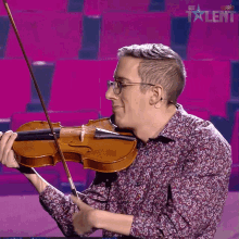 A person playing the violin performs on a stage with rows of empty red seats in the background. The person is wearing glasses and a patterned shirt