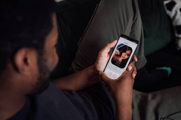 Person viewing a dating profile on a smartphone. The profile shows a woman's photo and interface elements