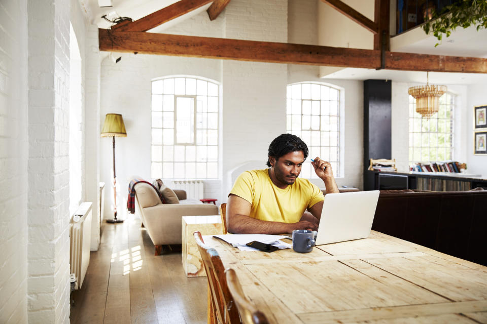 A guy wearing a yellow t-shirt works from home using his dining room table as a desk