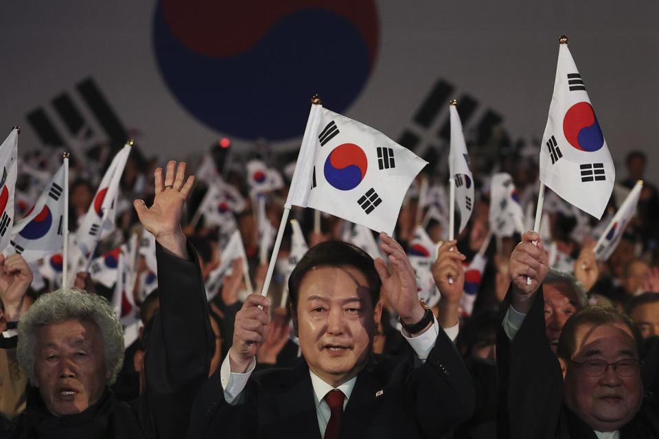 A man in dark suit and red tie holds a white, red and blue flag, surrounded by a crowd of people holding similar flags