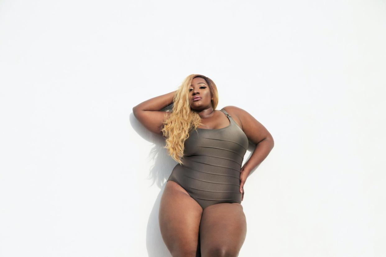 Black woman in a grey one piece swimsuit bathing suit swimwear posing in front of grey wall