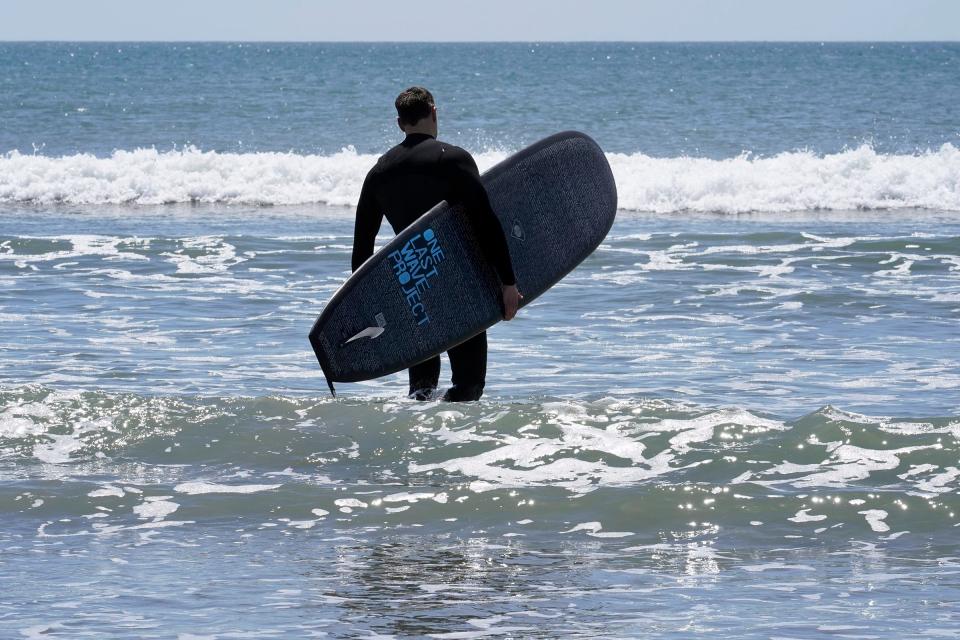 Dan Fischer, of Newport, R.I., walks into the ocean with his surfboard at Easton's Beach, in Newport, Wednesday, May 18, 2022. Fischer, 42, created the One Last Wave Project in January 2022 to use the healing power of the ocean to help families coping with a loss, as it helped him following the death of his father. Fischer places the names onto his surfboards, then takes the surfboards out into the ocean as a way to memorialize the lost loved ones in a place that was meaningful to them. (AP Photo/Steven Senne)