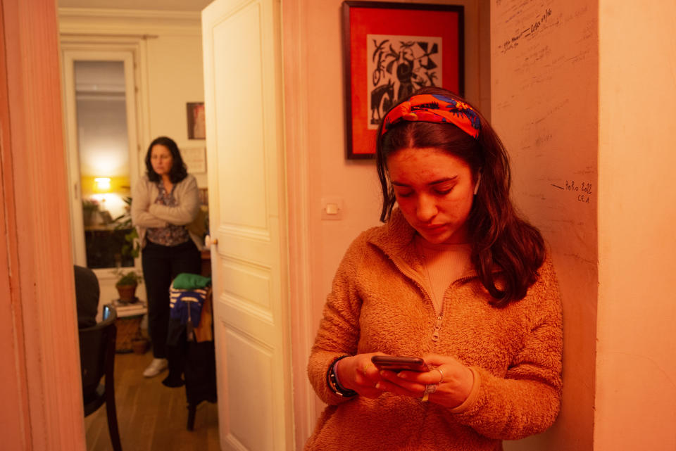 Anais Bulcao, 17, checks her phone on May 19, 2021, in the hallway of the Montmartre apartment where she lives with her parents, mother Nathalie Sartor, background, and father Joao Luiz Bulcao. Also living with them again is elder sister Livia Bulcao, who moved back in during the pandemic. As they grew up, the girls' heights were recorded with marks on the wall against which Anais is leaning. Nathalie says the pandemic distanced them from friends and "there are friends who live close by that we haven't seen, people from Montmartre, who are within walking distance." (AP Photo/Joao Luiz Bulcao)