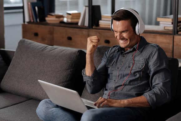 A man wearing headphones cheers as he looks at his laptop.