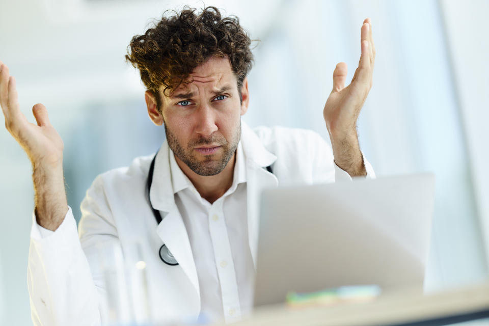 Man wearing a white coat with a stethoscope, sitting in front of a laptop with hands raised, looking frustrated