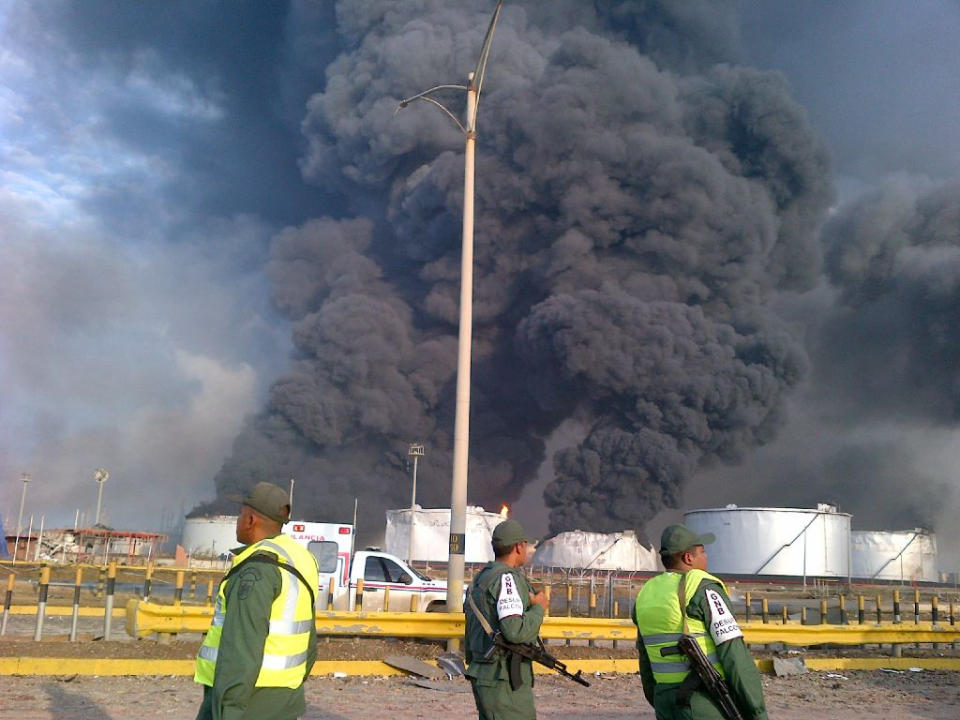 Large plumes of smoke rise from the Amuay refinery as national guards soldiers watch near Punto Fijo, Venezuela, Saturday, Aug. 25, 2012. A huge explosion rocked Venezuela's biggest oil refinery, killing and injuring dozens, an official said. (AP Photo/Abisaid Cermeno)