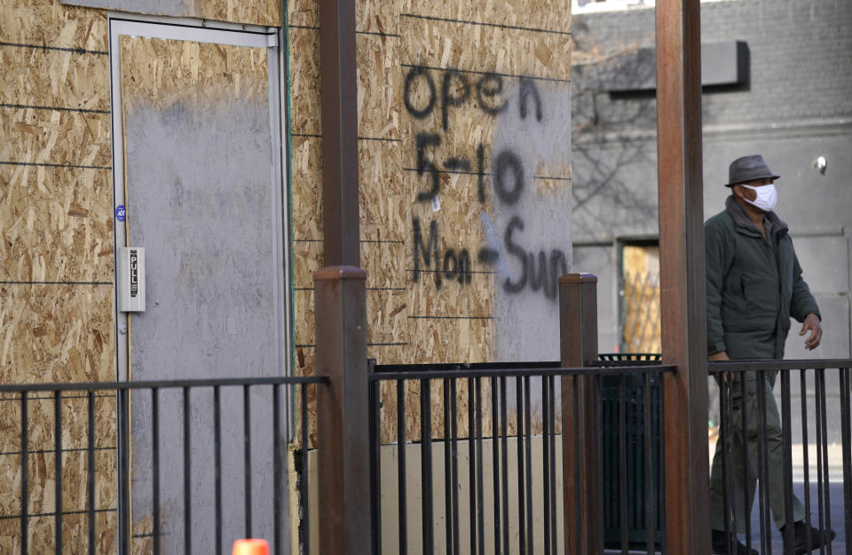 A pedestrain in a face mask walks past a restaurant with boarded-up windows on which hours of operation are listed along East Colfax Avenue Monday, Dec. 21, 2020, in downtown Denver. (AP Photo/David Zalubowski)