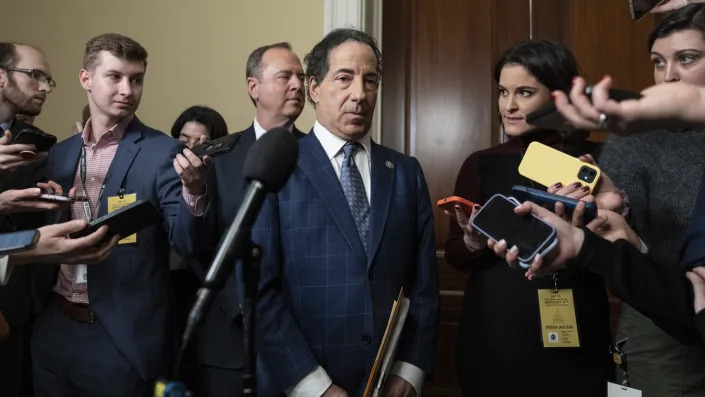 Rep. Jamie Raskin and Rep. Adam Schiff are surrounded by reporters with cellphones in hand, against a dark wood door.