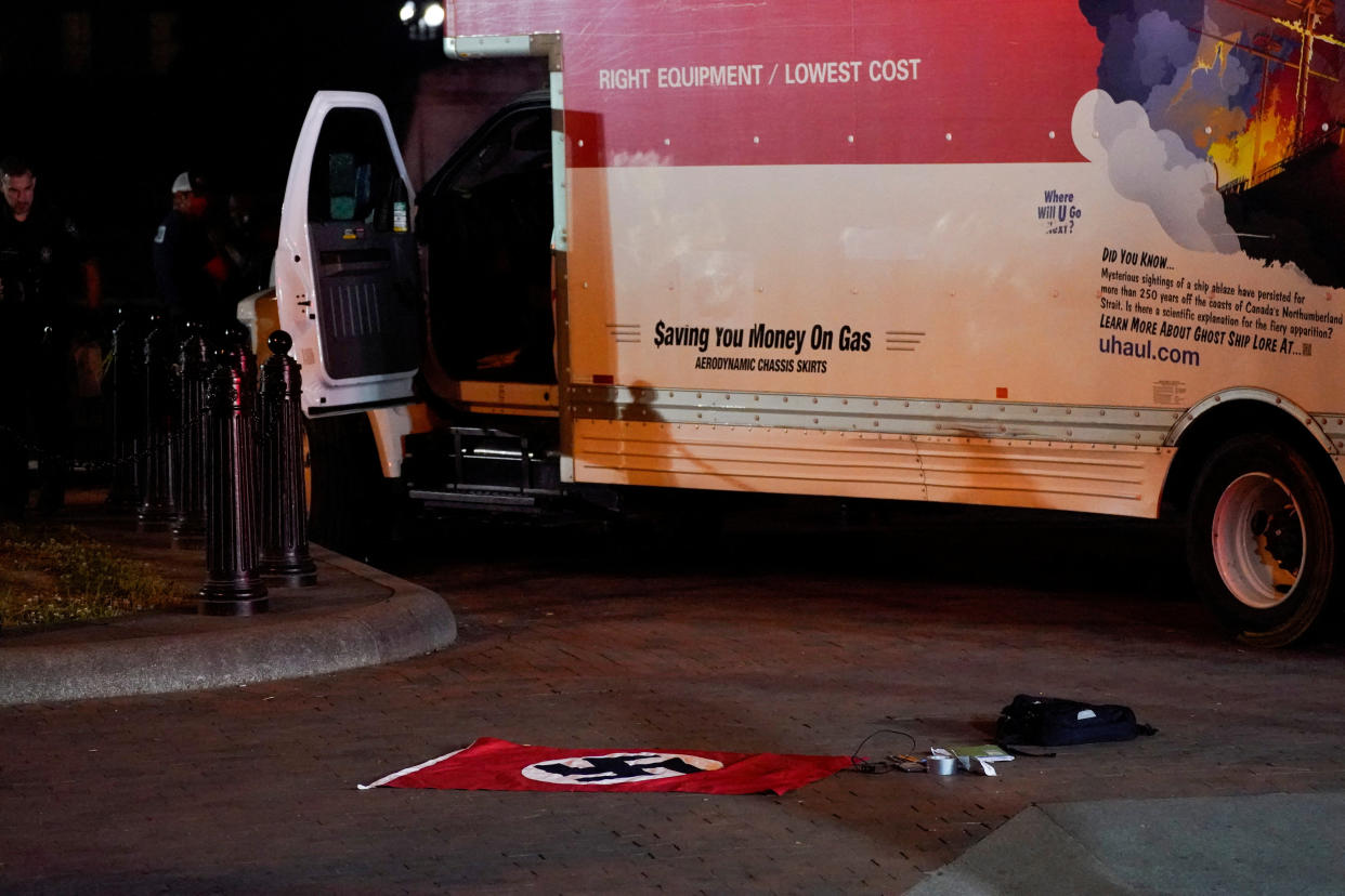 A flag emblazoned with a swastika and other objects lie in front of a Supermover U-Haul truck with its cab door open. Various slogans are printed on the sides of the truck, including: $aving You Money On Gas.