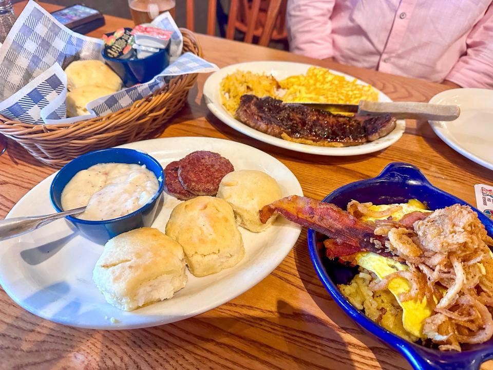 A wooden table filled with several plates, a bowl, and a basket of biscuits. A white plate with three biscuits, a plate with steak and eggs, and a bowl of eggs, hash browns, bacon, and fried onions sits on the table.