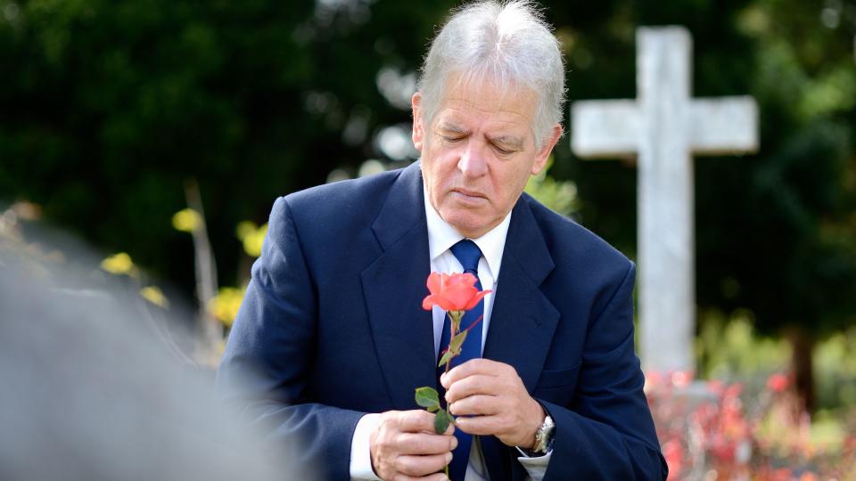 A grieving elderly man with a rose in a graveyard.