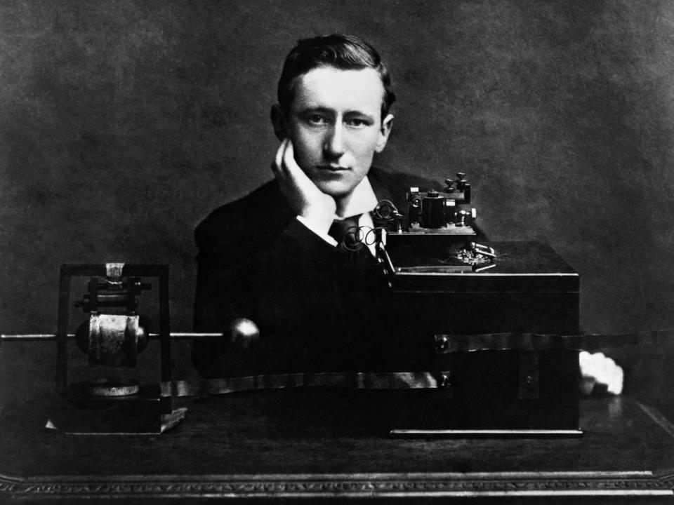 Gugliemo Marconi sitting with his hand under his chin while at a desk with the his electrical wireless apparatus