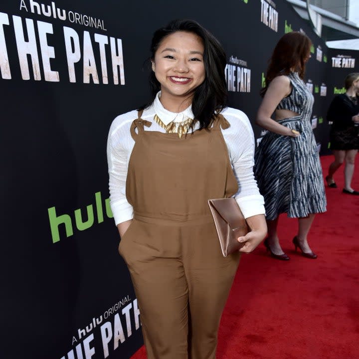 Hsu smiles on the red carpet wearing a jumpsuit and a long-sleeved dress shirt and a tiered necklace