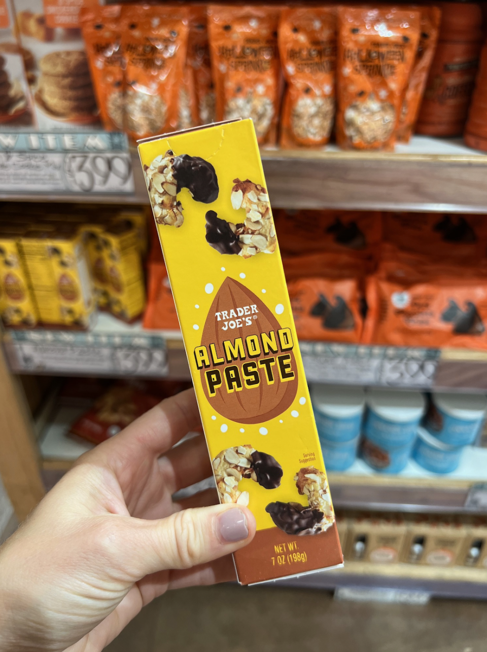 Hand holding a box of Trader Joe's Almond Paste in a grocery store aisle, with various other items visible on the shelves in the background