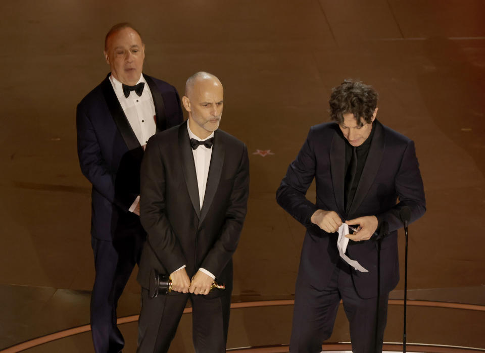 Three men on stage at an event, the central figure holding a paper, all in formal attire