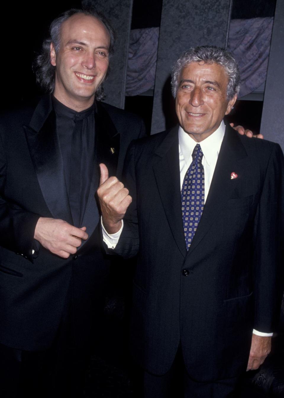 danny bennett and tony bennett stand next to each other and smile, tony raises a thumbs up toward danny, both men wear black suit jackets, tony also wears a blue patterned tie