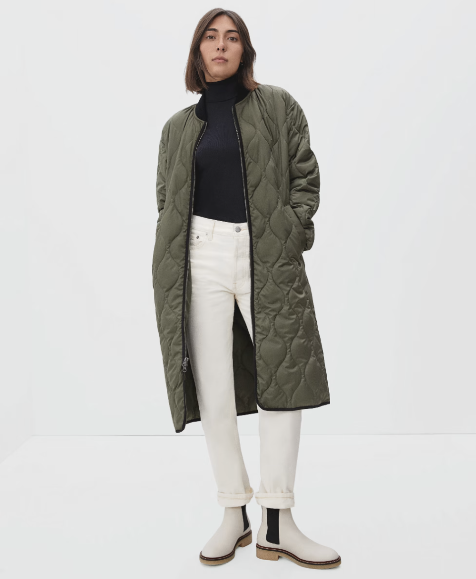 model wearing white boots, white jeans, black turtleneck and olive green The ReNew Long Liner in kalamata (photo via Everlane)