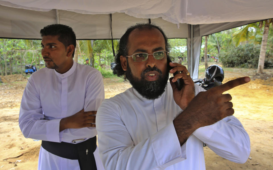 Sri Lankan Roman Catholic priests Father Niroshan Perera, right, speaks on his mobile phone with Father Anthony Nishan standing behind him at a mass burial ground for Easter Sunday's church explosion victims of Katuwapitiya in Colombo, Sri Lanka, Thursday, April 25, 2019. Christians in Sri Lanka belong to both its main ethnic groups, and that rare inclusiveness of a small religious minority may explain the measured calm that’s been the response so far to the Easter attacks. But there’s widespread fear that more attacks could plunge Sri Lanka into the cycle of violence and retaliation that marked the bloody civil war that ended a decade ago. (AP Photo/Manish Swarup)