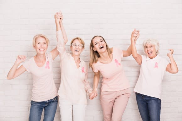 Four women wearing pink breast cancer ribbons standing against a white brick wall and smiling with arms raised.