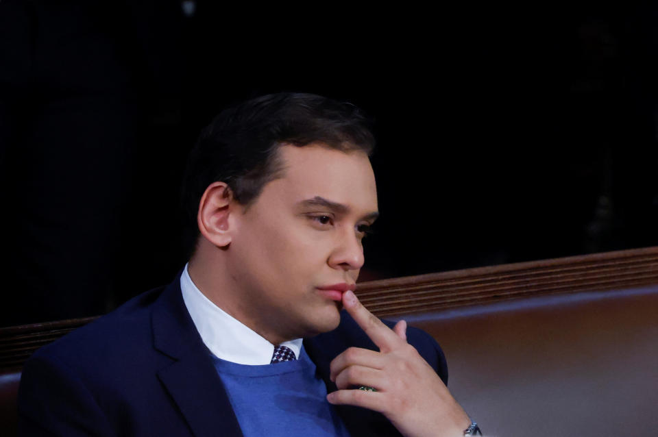 Rep. George Santos in crisp shirt and jacket, lifts his index finger to his lips in the House Chamber.