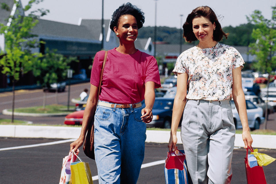 Two women walk in a parking lot carrying shopping bags. One wears a pink shirt and jeans, while the other wears a floral blouse and gray pants