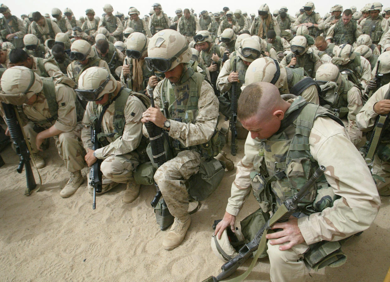 Serried ranks of Marines, their rifles pointed down into the sand, kneel and pray. All but the Marine in front are wearing helmets, and his hair is shaved into a tonsure..