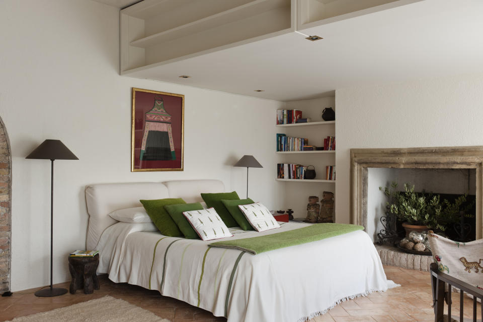 A cozy bedroom features a neatly made bed with green accent pillows, a bookshelf, a stone fireplace, a piece of artwork above the bed, and floor lamps