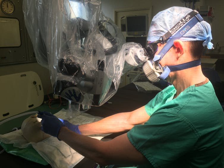 A surgeon in scrubs and a hairnet wears googles and a respirator mask and looks down a microscope while simulating an operation on a skull.