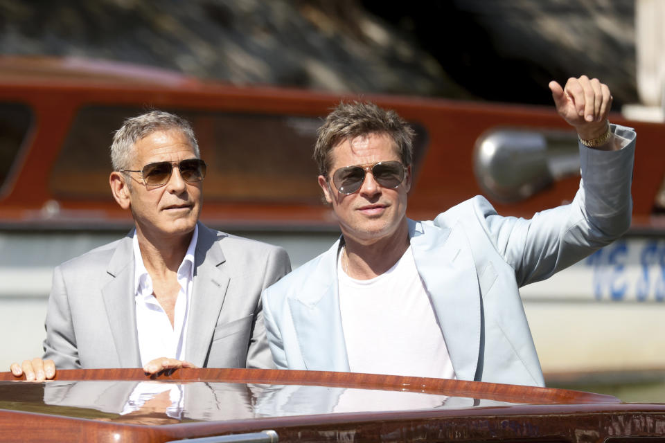 George Clooney and Brad Pitt on a boat; Clooney in a grey suit and sunglasses, Pitt in a light jacket and sunglasses, waving