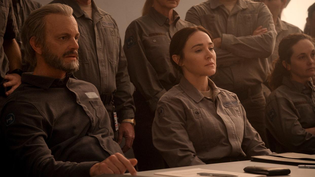  Sitting at a table are three people with very serious expressions. In the background there are more people standing, some with their arms crossed. They are all wearing the same gray jumpsuit. 